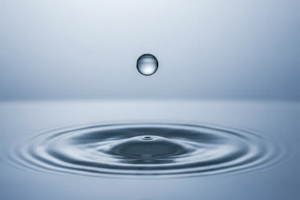 Single water droplet above calm surface creating concentric ripples on a soft blue-gray background.