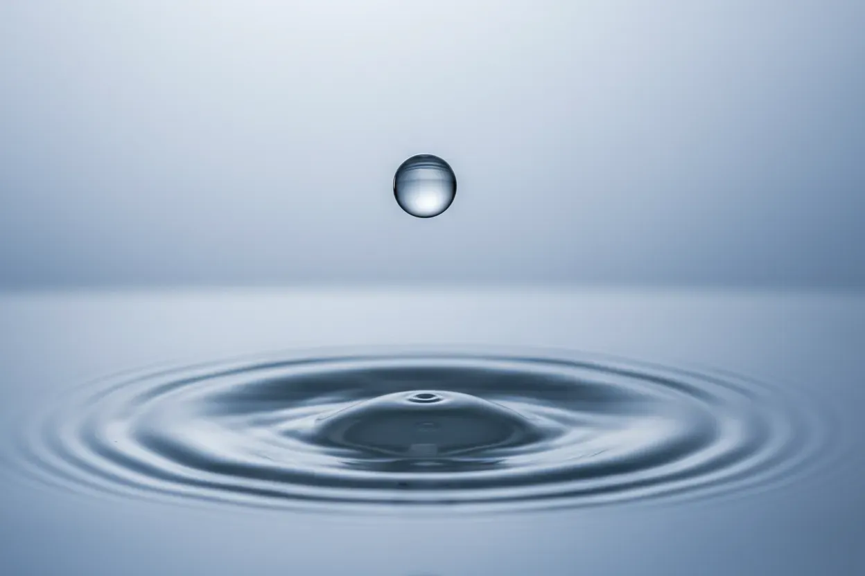 Single water droplet above calm surface creating concentric ripples on a soft blue-gray background.